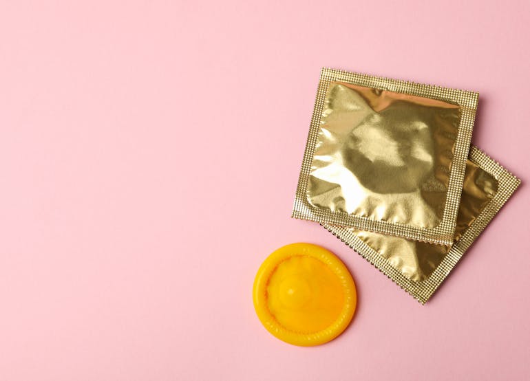Image of square gold foil packaged condoms on a pink background, with one orange condom outside of the packaging unopened as a disc
